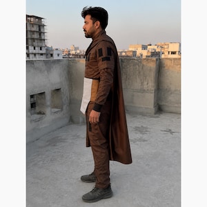 Puede incluir: Un atuendo marr&oacute;n con capa, que consta de una chaqueta, pantalones y un cintur&oacute;n blanco y beige. El atuendo lo lleva una persona de pie sobre una superficie de hormig&oacute;n con edificios al fondo.