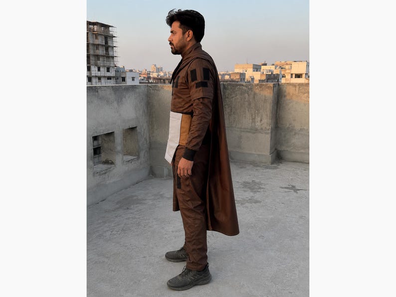 Puede incluir: Un atuendo marr&oacute;n con capa, que consta de una chaqueta, pantalones y un cintur&oacute;n blanco y beige. El atuendo lo lleva una persona de pie sobre una superficie de hormig&oacute;n con edificios al fondo.