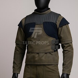 May include: Olive green and navy blue tactical vest with the text "TASTIC PROPS". The vest has a padded collar and shoulder accents, with two black buckles on the front. The long-sleeved shirt underneath is olive green.