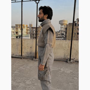 Pu&ograve; includere: Una persona indossa una tuta grigia con collo alto e gilet beige. L'outfit ha un design futuristico, con una tasca sulla gamba. Lo sfondo mostra un paesaggio urbano e un serbatoio d'acqua.