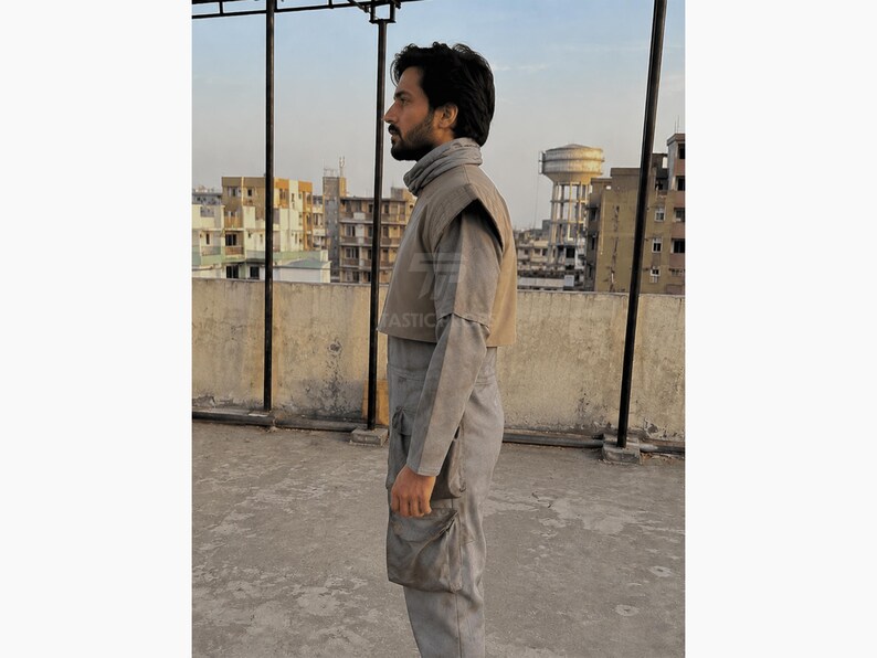 Pu&ograve; includere: Una persona indossa una tuta grigia con collo alto e gilet beige. L'outfit ha un design futuristico, con una tasca sulla gamba. Lo sfondo mostra un paesaggio urbano e un serbatoio d'acqua.