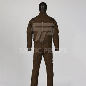 Pu&ograve; includere: Un costume marrone esposto su un manichino. L'outfit include una giacca con cerniera e spalline, e pantaloni abbinati. La giacca ha cuciture geometriche sulle maniche. Il manichino ha una testa nera.