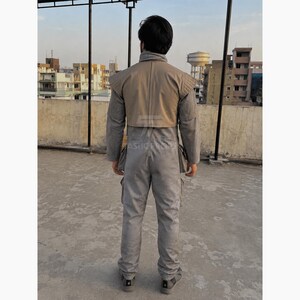 Pu&ograve; includere: Tuta grigia con spalla color beige. La tuta ha pi&ugrave; tasche e un colletto alto. La persona &egrave; in piedi su una superficie di cemento con edifici sullo sfondo.