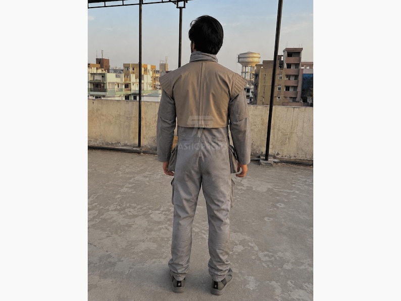 Pu&ograve; includere: Tuta grigia con spalla color beige. La tuta ha pi&ugrave; tasche e un colletto alto. La persona &egrave; in piedi su una superficie di cemento con edifici sullo sfondo.