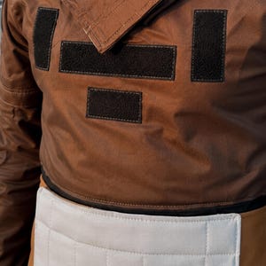 Puede incluir: Chaqueta marr&oacute;n con parches rectangulares negros y un panel acolchado blanco en la cintura. La chaqueta tiene cuello y parece estar hecha de un material duradero. El dise&ntilde;o sugiere una est&eacute;tica utilitaria o de ropa de trabajo.