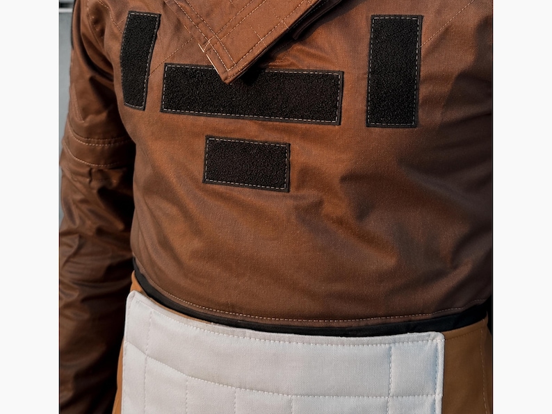 Puede incluir: Chaqueta marr&oacute;n con parches rectangulares negros y un panel acolchado blanco en la cintura. La chaqueta tiene cuello y parece estar hecha de un material duradero. El dise&ntilde;o sugiere una est&eacute;tica utilitaria o de ropa de trabajo.