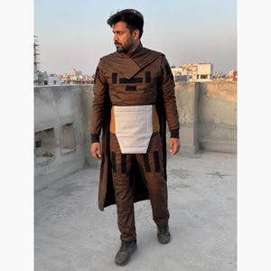 Puede incluir: Un disfraz marr&oacute;n con detalles negros, que incluye un abrigo largo, pantalones y una pieza para el pecho. El atuendo tiene un dise&ntilde;o futurista, con un cuello alto y elementos en capas. La persona lleva zapatos oscuros y est&aacute; de pie en una azotea con un fondo de ciudad.