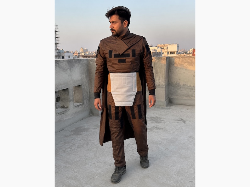 Puede incluir: Un disfraz marr&oacute;n con detalles negros, que incluye un abrigo largo, pantalones y una pieza para el pecho. El atuendo tiene un dise&ntilde;o futurista, con un cuello alto y elementos en capas. La persona lleva zapatos oscuros y est&aacute; de pie en una azotea con un fondo de ciudad.