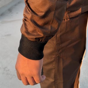 Puede incluir: Primer plano de una manga de chaqueta marr&oacute;n con pu&ntilde;o acanalado negro. La chaqueta parece estar hecha de un material liso, posiblemente resistente al agua. La mano es visible, con los dedos ligeramente curvados.