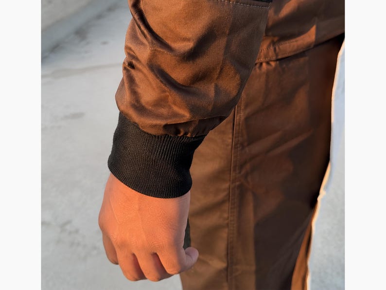 Puede incluir: Primer plano de una manga de chaqueta marr&oacute;n con pu&ntilde;o acanalado negro. La chaqueta parece estar hecha de un material liso, posiblemente resistente al agua. La mano es visible, con los dedos ligeramente curvados.