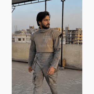 Pu&ograve; includere: Tuta grigio chiaro con collo alto e spalline. La tuta ha pi&ugrave; tasche e un design aderente. Il testo "TASTIC PROPS" &egrave; visibile sul petto. L'outfit &egrave; indossato all'aperto.