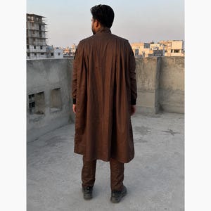 Puede incluir: Una persona con un abrigo largo marr&oacute;n y pantalones a juego. El abrigo tiene una parte delantera abotonada y un dise&ntilde;o largo y fluido. La persona est&aacute; de pie sobre una superficie de hormig&oacute;n con edificios al fondo.