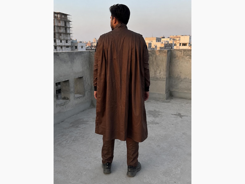 Puede incluir: Una persona con un abrigo largo marr&oacute;n y pantalones a juego. El abrigo tiene una parte delantera abotonada y un dise&ntilde;o largo y fluido. La persona est&aacute; de pie sobre una superficie de hormig&oacute;n con edificios al fondo.