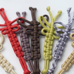 May include: A collection of six paracord keychains featuring intricate knot work in red, dark brown, light brown, lime green, purple, and a mottled grey and white pattern. Each keychain is crafted with a central braided design and loops.
