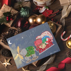 Op de afbeelding: Een kerstkaart met een olifant die een slee vol cadeaus trekt, tegen een sterrenhemel. De kaart is omgeven door ingepakte cadeaus, kerstballen, dennenappels en linten op een houten oppervlak.