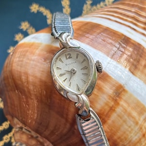 May include: Vintage Bulova watch with a silver-toned case and a cream-colored face. The watch features a small, oval-shaped dial with silver hands and hour markers. The watch band is a silver-toned, flexible design. The watch is displayed on a seashell.