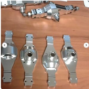 以下が含まれることがあります： 光沢のある銀色の金属機械部品のコレクション。おそらく車両または機械用。部品には、丸みを帯びた長方形のセクションを持つ細長い部品と、より複雑に組み立てられたコンポーネントが含まれます。木製の表面に配置されています。