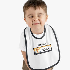 May include: White baby bib with a black trim and the text "MY NAME IS" above a name tag that says "NOAH" with a giraffe graphic. The bib is worn by a smiling toddler.