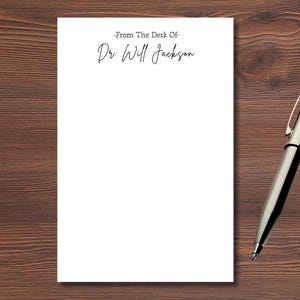 May include: A white notepad with the text "-From The Desk Of- Dr Will Jackson" and a silver pen on a dark brown wooden surface. The pen is positioned diagonally across the right side of the notepad.