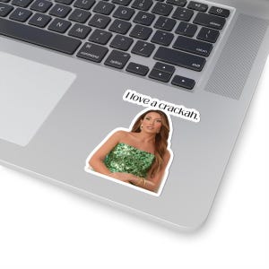 Peut inclure: Un autocollant en vinyle découpé représentant une personne aux longs cheveux bruns portant un haut bustier à sequins verts et des bracelets dorés. Au-dessus, le texte I love a crackah est écrit en police serif noire sur un ordinateur portable.