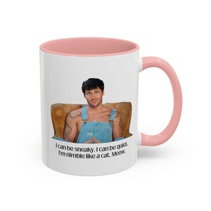 Op de afbeelding: Witte keramische mok met een roze binnenkant en handvat. De mok heeft een afbeelding van een persoon en de tekst "I can be sneaky. I can be quiet. I'm nimble like a cat. Meow."