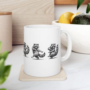 Puede incluir: Taza de cerámica blanca con una ilustración en blanco y negro de tres mapaches bailando. Los mapaches se muestran en diferentes poses, con sus colas rayadas y máscaras faciales. La taza tiene un asa curva.