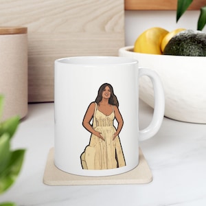 Puede incluir: Taza de cerámica blanca con una ilustración de dibujos animados de una persona con un vestido amarillo. La ilustración tiene un contorno negro. La taza está sobre un posavasos beige.