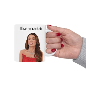 Può includere: Tazza in ceramica bianca con un'illustrazione a cartoni animati di una donna con un vestito rosso e una collana d'argento. La tazza ha il testo "I love a crackah." stampato in nero. La tazza è tenuta da una mano con lo smalto rosso.