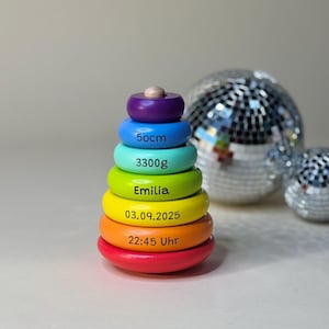Puede incluir: Un juguete apilable de madera de colores del arcoíris, con una parte superior morada y anillos azules, verdes, amarillos, naranjas y rojos. Los anillos están inscritos con: 50cm, 3300g, Emilia, 03.09.2025 y 22:45 Uhr. Una pequeña bola de madera está encima.