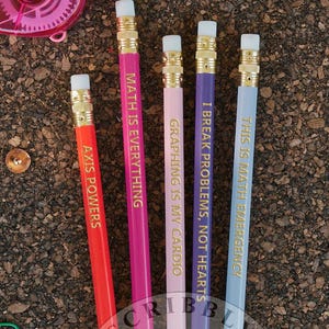 Puede incluir: Un conjunto de lápices coloridos con citas motivadoras, junto con clips, chinchetas, una cinta correctora rosa y un pequeño bloc de notas sobre una superficie marrón texturizada. Los lápices son rojos, rosas, morados, lavanda y azul claro.