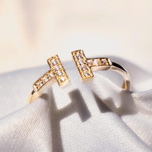 Puede incluir: Un anillo abierto de color dorado con dos extremos en forma de T, cada uno incrustado con pequeñas piedras preciosas transparentes. El anillo se presenta sobre un fondo de tela blanca y suave, creando un contraste que resalta el diseño y el brillo del anillo.