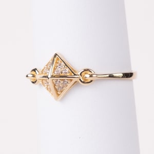 May include: A gold-colored ring featuring a geometric diamond-shaped design. The ring's center is adorned with small, sparkling stones. The ring is displayed on a white background, showcasing its intricate details and craftsmanship.