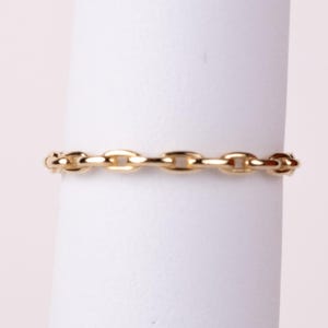 May include: A gold ring featuring a continuous chain link design. The band is composed of small, uniform, polished oval links connected in a series, creating a flexible-looking appearance with a bright, reflective metallic finish throughout.