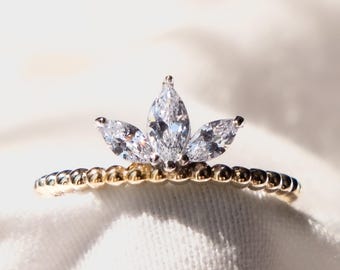 Puede incluir: Un anillo de oro con un diseño de racimo de diamantes talla marquesa. El anillo presenta una banda con cuentas y una disposición de tres piedras de diamantes claros y brillantes, creando una apariencia floral o de corona.