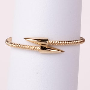 May include: A gold-toned bypass ring featuring a twisted rope-textured band that tapers into two smooth, pointed arrow-like ends that overlap at the center. The metallic surface has a polished finish, creating a reflective appearance on the points.