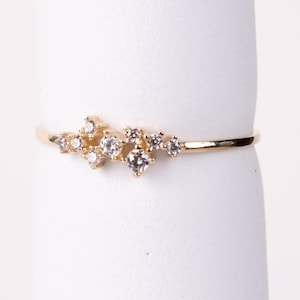 May include: A delicate gold ring featuring a cluster of small, sparkling clear gemstones. The ring's band is thin and polished, and the gemstones are arranged in a flowing, asymmetrical design. This elegant ring is perfect for everyday wear or special occasions.