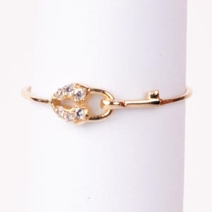 May include: A delicate gold-tone ring featuring a key and lock design. The lock portion is embellished with small, clear gemstones. The ring is set against a white background, highlighting its intricate details and elegant style.