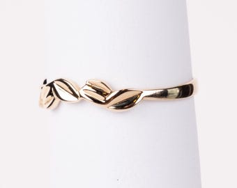 Puede incluir: Un anillo dorado con un delicado diseño de hojas. El anillo tiene una banda lisa y hojas texturizadas. Las hojas están dispuestas en un patrón repetitivo alrededor del anillo. El anillo se muestra sobre un fondo blanco. Joyería.