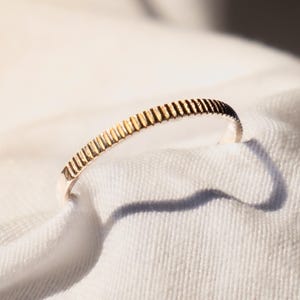Puede incluir: Un anillo dorado con un diseño texturizado y acanalado. El anillo está sobre un fondo de tela blanca, con sombras proyectadas por la luz solar. La superficie del anillo tiene una serie de líneas verticales, creando un patrón único. Es una joya.
