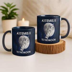 Peut inclure: Deux mugs en céramique bleu marine avec du texte blanc et un motif de fusée. Les tasses présentent le texte "ARTEMIS II" et "TO THE MOON" avec un graphique de pleine lune. Un mug est sur un dessous de verre en bois.