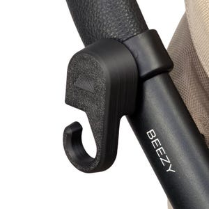 May include: Close-up of a black stroller handle with the word "BEEZY" printed in white. The handle has a curved, textured grip and a black hook attachment. A beige fabric is visible in the background.