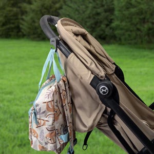 May include: A tan stroller with a black handle and a matching tan canopy. A light blue and tan backpack with a sloth pattern is hanging from the handle. The stroller is on a green grassy background.