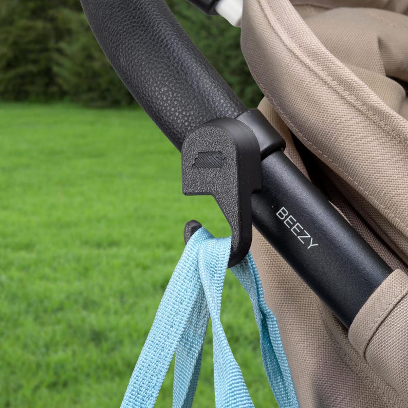 May include: A black stroller hook attached to a black handle with the word "Beezy" printed on it. A light blue strap is hanging from the hook. The background is a blurred green lawn.