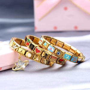 Puede incluir: Tres pulseras de dijes doradas con dijes de esmalte coloridos. Los dijes presentan varios diseños, incluyendo corazones, estrellas y notas musicales. Una pulsera tiene un dije en forma de estrella con una perla. Las pulseras se exhiben sobre una superficie rosa claro.