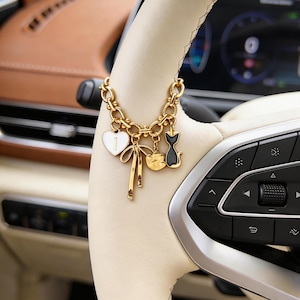 May include: Gold-coloured car charm with a chain and various charms. Charms include a white heart with the number 1, a bow, a cat, and a flower. The charm is hanging on a cream-coloured steering wheel.