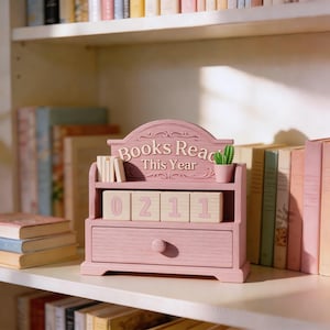 Puede incluir: Un organizador de escritorio de madera rosa con forma de estanter&iacute;a, con el texto "Books Read This Year". Tiene un caj&oacute;n peque&ntilde;o, una estanter&iacute;a con libros peque&ntilde;os y bloques de madera que muestran "0211". Un peque&ntilde;o cactus en maceta est&aacute; encima.