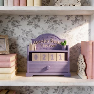 Puede incluir: Un organizador de madera lila con forma de estanter&iacute;a con el texto "Books Read This Year". Tiene un caj&oacute;n y ranuras para bloques de n&uacute;meros de madera que muestran "0211". Un peque&ntilde;o cactus se encuentra en la parte superior, con una pila de libros a la izquierda.