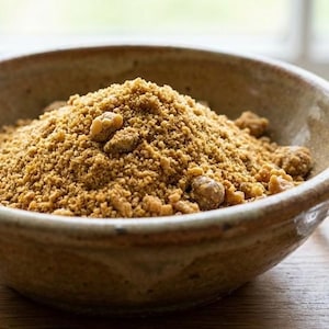 May include: A ceramic bowl filled with brown sugar, with a spoon resting on the side. The sugar is a light brown color and has a granular texture. The bowl is a light brown color with a slightly rough texture.
