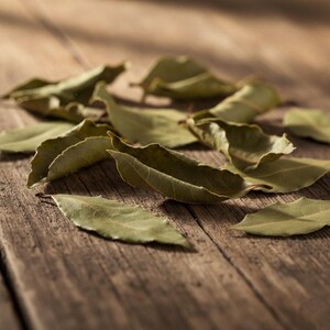 May include: A collection of dried bay leaves, with a muted green hue, scattered on a weathered wooden surface. The leaves display a natural, organic texture, with visible veins and slightly curled edges. The lighting highlights the leaves' details.
