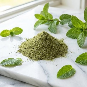 May include: A pile of green powder and fresh mint leaves on a white marble surface. The mint leaves are bright green and arranged around the powder, with some sprigs and individual leaves scattered about. The scene is lit by natural light.